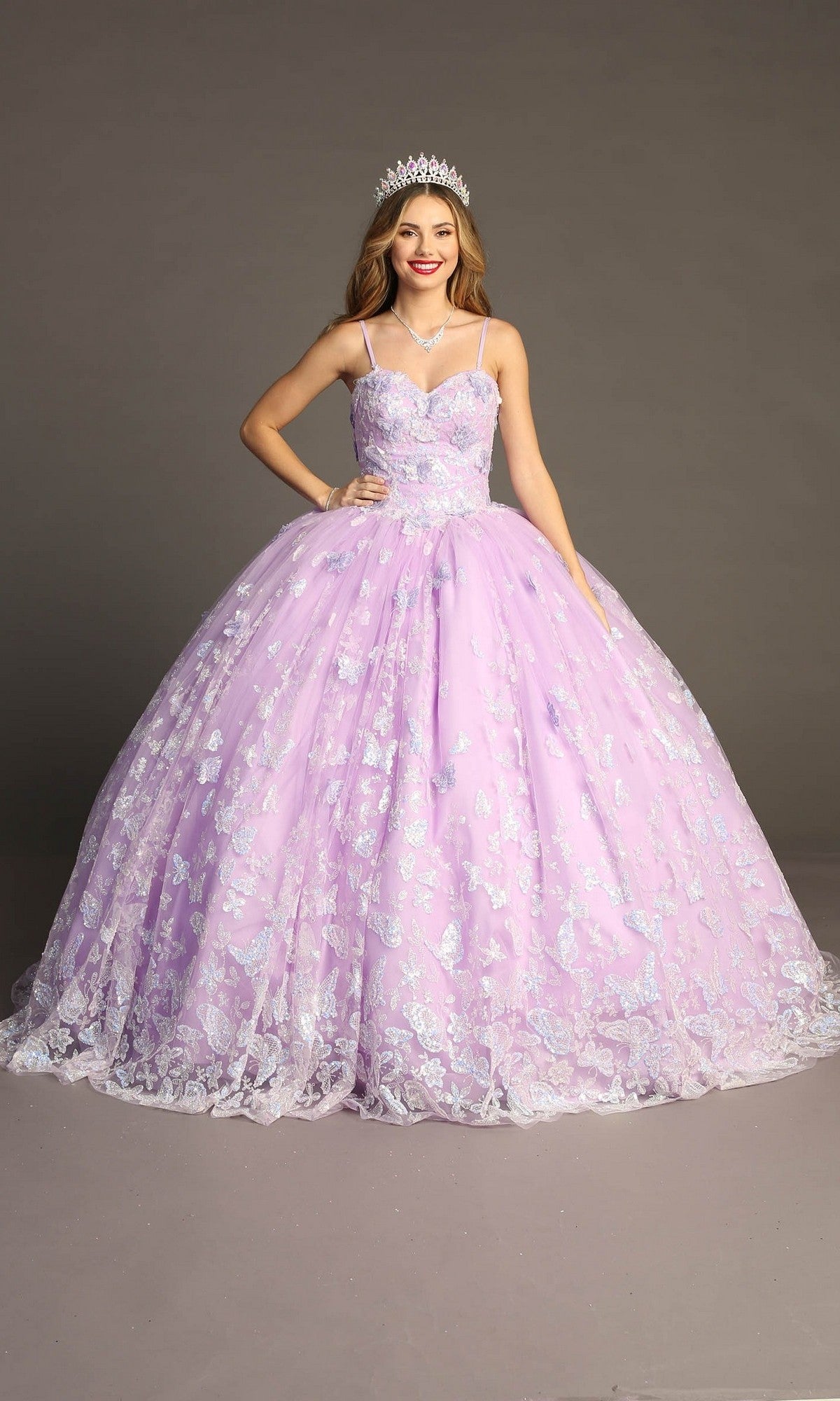 A young woman wearing the Quinceanera Dress CHF3097 By Chicas, featuring sparkly lilac glitter tulle and delicate butterfly appliques, paired with a silver tiara and red lipstick, stands against a plain gray background.